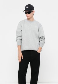 Grijze sweatshirt met ronde halslijn, zwarte broek, zwarte pet en zonnebril. De sweatshirt heeft een klein logo op de borst.
