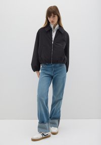 Schwarze Bomberjacke mit Reißverschluss, ausgestattet mit einem spitzen Kragen und gerippten Bündchen, kombiniert mit hellblauen gerade geschnittenen Jeans und weißen Sneakers.
