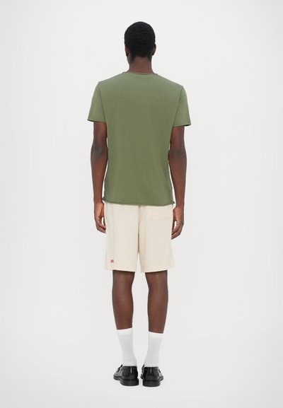 Green short-sleeve t-shirt with a crew neck, paired with beige shorts featuring a subtle pattern and small red branding. Black shoes and white socks.