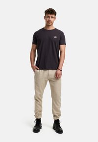 Graues Baumwoll-T-Shirt mit einem kleinen weißen Grafikdesign, kombiniert mit hellbeigen Leinen-Jogginghosen und schwarzen Schnürstiefeln. Das Modell steht mit den Händen in den Taschen.