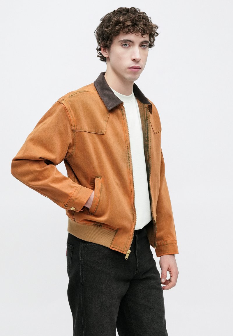 Brown corduroy jacket with a dark collar, zip front, and pockets. Worn over a light colored shirt, paired with black pants.