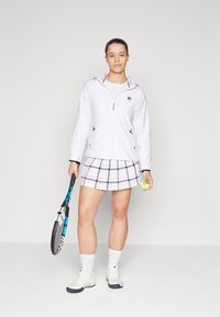 Hvit zip-up jakke med hette, rutete skjørt i hvit, marineblå og rosa, hvite tennissko, holder en tennisracket og ball.