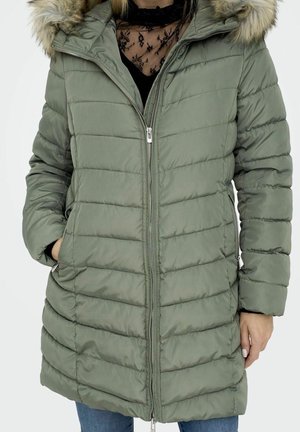 Femme portant un manteau d'hiver vert matelassé avec une fermeture éclair et une capuche bordée de fourrure, les mains dans les poches latérales, associée à un jean bleu.