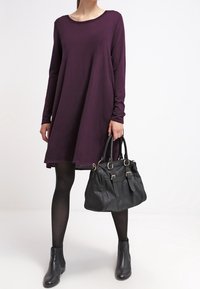Robe mauve à manches longues, longueur au-dessus du genou, associée à des bottines noires à la cheville et à un sac à main en cuir noir avec des détails métalliques et une forme structurée.