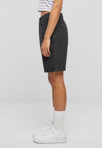 Shorts en denim noir avec une coupe droite, taille mi-haute, et un design à cinq poches, associés à des chaussures de sport blanches et des chaussettes.