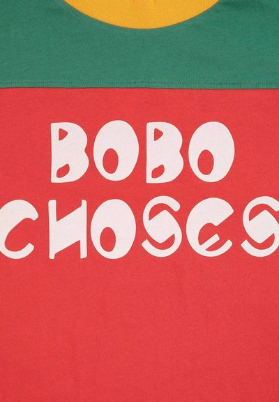 Červené bavlnené tričko s veľkým bielym textom "BOBO CHOSES", umiestnené na zelenom jho a žltom kruhovom akcentu navrchu.