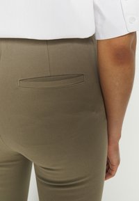 Calças verde-oliva com uma textura suave, apresentando um único bolso traseiro e detalhes de costura discretos. Combinadas com uma camisa branca.