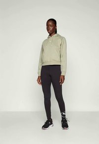 Sudadera verde-azeitona com cordão e punhos canelados, combinada com leggings ajustadas pretas e sapatilhas desportivas pretas com detalhes em rosa.