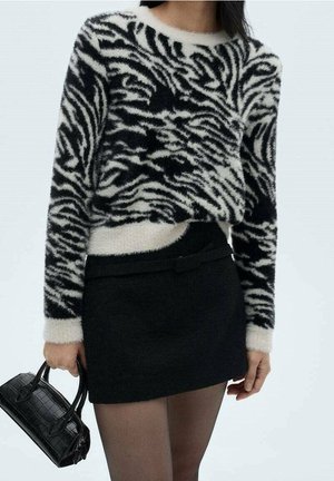 Kvinde iført en sort og hvid zebra-mønstret sweater i blødt materiale, sort mininederdel, gennemsigtige sorte strømpebukser og holder en lille sort håndtaske.