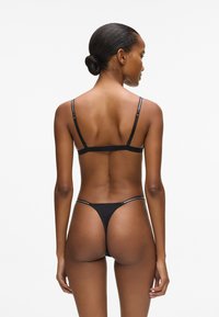 Juodas apatinis komplektas, kuriame yra plonų dirželių liemenėlė ir stringai. Lygi medžiaga, minimalistinis dizainas, reguliuojami dirželiai ir subtilus logotipo detalė.