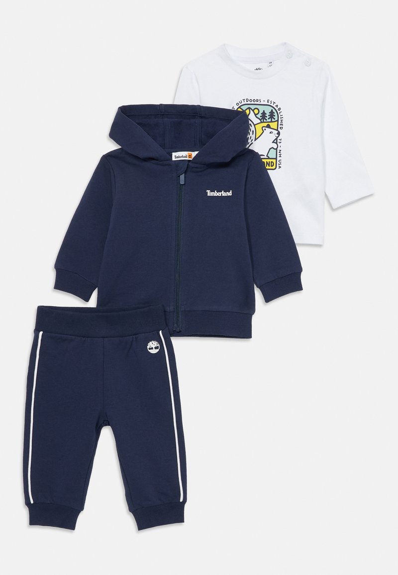 Conjunto infantil de três peças composto por um hoodie azul-marinho com fecho éclair, uma camisa de manga longa branca com um logótipo e calças azul-marinho com riscas laterais.