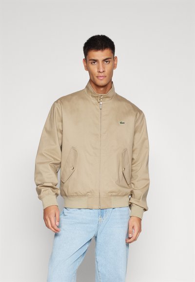 Veste bomber beige en tissu lisse, dotée d'une fermeture éclair, d'un col montant et de poches latérales avec rabats. Logo vert sur la poitrine.