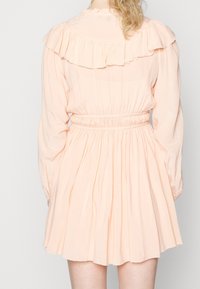 Pfirsichfarbenes Kleid mit Rüschenausschnitt, langen Ärmeln, elastischem Taillenbund und weitem Rock. Weicher Stoff, schlichte Textur, knielang.