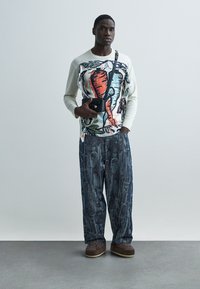 Καθημερινό σύνολο που περιλαμβάνει ένα ελαφρύ πουκάμισο με μακριά μανίκια και πολύχρωμο μοτίβο, καθώς και φαρδιά παντελόνια με μπλε σχέδιο. Αξεσουάρ περιλαμβάνουν μια μικρή μαύρη τσάντα.