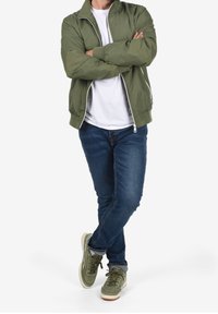 Bomber jacket verde oliva con zip, t-shirt bianca, jeans blu e sneakers verdi. L'outfit presenta una vestibilità rilassata e uno stile informale.