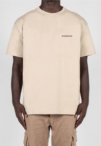 Béžové bavlněné tričko s krátkým rukávem, kulatým výstřihem a malým černým textem "MJONZALES" na hrudi; uvolněný střih a hladká textura.