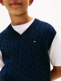 Tamno plavi pletivi prsluk s V-izrezom, nosi se preko bijelog T-shirt-a; ima malo oznaku uz vrat.