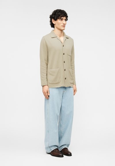 Ανοιχτό μπεζ πουκάμισο-τζάκετ με μακριά μανίκια, μπροστινή τσέπη και κούμπωμα με κουμπιά, σε συνδυασμό με άνετο, ανοιχτό μπλε τζιν και σκούρα παπούτσια.