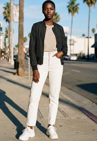 Staande persoon die een zwart bomberjack, een beige shirt, witte broek met hoge taille en witte sneakers draagt op een zonnige stedelijke straat.