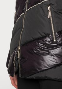 Giacca imbottita nera e viola scuro, con sezioni di tessuto lucido e opaco, cerniere metalliche e dettagli di cucitura orizzontali.