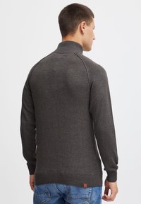 Mørkegrå høyhalset genser med ribbestrikkede mansjetter og nederkant, med teksturert strikkemønster, brukt over lyseblå jeans. Brun logo nær nederkanten.