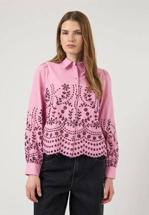 Žena v růžové halence s knoflíky, tmavým květinovým vyšíváním a nafouknutými rukávy, v černých džínách, na jednoduchém světlém pozadí.