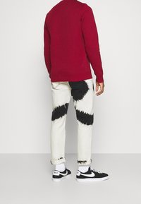 Sweat-shirt rouge avec poignets et ourlet côtelés, associé à un jean tie-dye blanc avec motifs noirs, et des baskets noires Nike.
