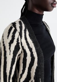 Kraujantinis kardiganas su juodais ir kremiškai baltais zebra raštais, pasižymintis minkšta tekstūra ir atvira priekiu, dėvimas ant juodo golfuko.