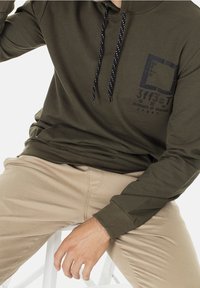 Olijfgroen hoodie met een voorzak, zwart-witte getailleerde koorden en een grafische print op de borst. Gecombineerd met beige broek.