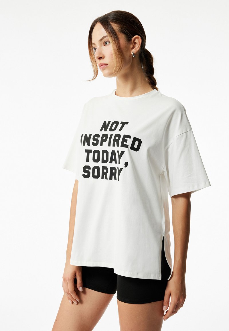 Valkoinen puuvillapaita, jossa mustalla tekstillä lukee "EI INSPIROITUNUT TÄNÄÄN, PÄIVÄN SUOSIO." Miellyttävä istuvuus ja sivuhalkiot. Yhdistetty mustiin shortseihin.