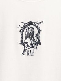 Črna vezenina skice šivanja punčke, ki drži cvet, obkrožena s prepletenimi vinsko trtami, s tekstom "GAP" spodaj na beli tkanini.