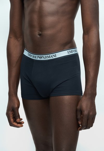 Αρσενικός κορμός που φοράει μαύρα μποξεράκια Emporio Armani με αντίθετη λευκή ζώνη μέσης και λογότυπο, στέκεται μπροστά σε φωτεινό φόντο.