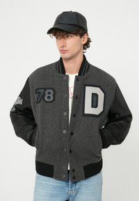 Mladý muž v sivom a čiernom študentskom kabáte s nášivkami „78“ a „D“, čiernou čiapkou, bielou košeľou, svetlomodrými džínami, ruky v vreckách.