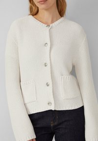 Cardigan en tricot blanc avec un col rond, cinq boutons en argent, deux poches avant et des poignet côtelés. Tissu texturé et design court.