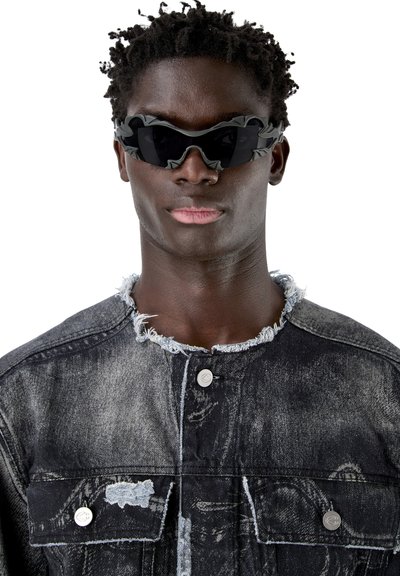 Man met kort krullend haar, die zwarte sculpturale zonnebrillen draagt en een zwarte acid-wash spijkerjas met rafelige kraag en zakken.