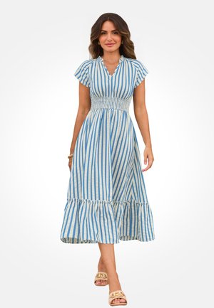Femme portant une robe midi à rayures verticales bleues et blanches avec une taille smockée et un ourlet volanté, marchant vers l'avant sur un fond blanc.