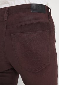 Burgundské džíny z denimu s matnou texturou, s černou koženou visačkou a zadními kapsami s viditelnými švy.