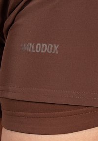 Bruna sportshorts tillverkade av slät, stretchig tyg, med en synlig logotyp "SMILODOX" i en något upphöjd textur.