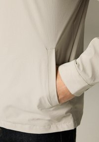 Beżowa lekka kurtka o gładkiej fakturze, z boczną kieszenią i mankietami. Projekt jest minimalistyczny i nowoczesny.
