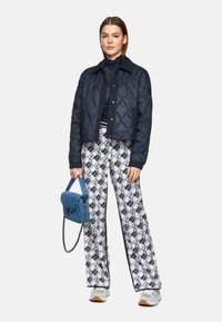 Gepolsterte marineblaue Jacke, schwarzes Spitzenoberteil, gemusterte weiße Hose mit blauen Akzenten und eine blaue gepolsterte Handtasche. Das Model trägt helle Turnschuhe.