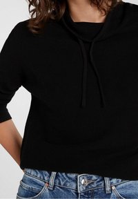 Svart koftatröja med kappa och trekvartslånga ärmar, med dragsko och strukturerad tyg. Matchad med blå denimjeans.