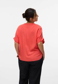Blusa de manga corta de coral con puños enrollados, corte relajado, escote redondeado y textura suave, combinada con pantalones de mezclilla oscuros.