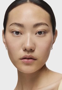 Primer plano de un rostro con piel suave, ojos oscuros y un maquillaje sutil. Lleva pendientes de plata pequeños y circulares, con un fondo neutro.