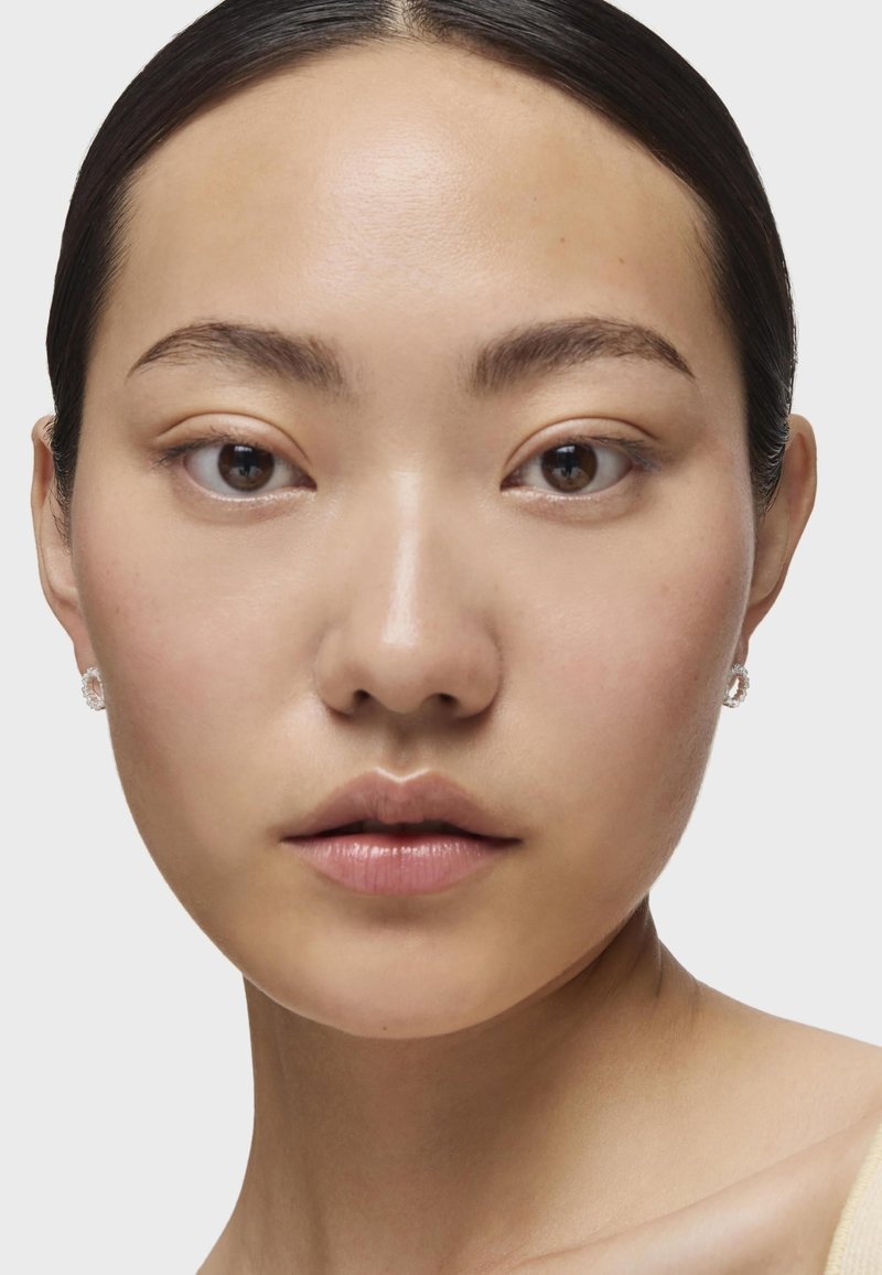 Primer plano de un rostro con piel suave, ojos oscuros y un maquillaje sutil. Lleva pendientes de plata pequeños y circulares, con un fondo neutro.