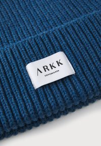 Bonnet en tricot côtelé bleu avec une étiquette blanche sur laquelle est imprimé en noir "ARKK COPENHAGEN". La texture est épaisse et confortable, offrant une chaleur optimale.