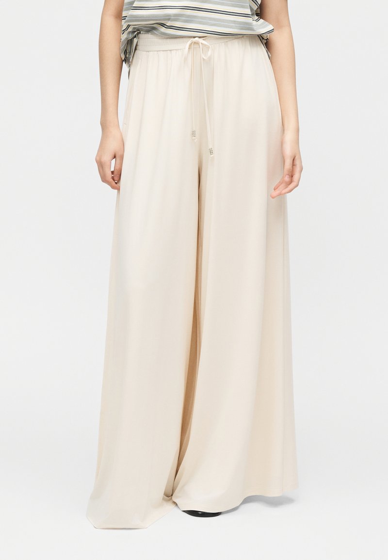 Μπεζ παντελόνι φαρδιάς γραμμής με κορδόνι στη μέση, συνδυασμένο με ριγέ πουκάμισο μπηγμένο μέσα, εμφανίζοντας μόνο τον κάτω κορμό και τα πόδια.