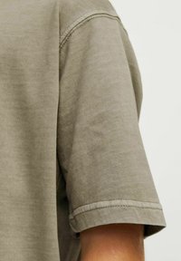 T-shirt s krátkým rukávem v olivově zelené barvě, vyrobený z měkké, texturované látky s volným střihem. Viditelné švy na lemu rukávu.