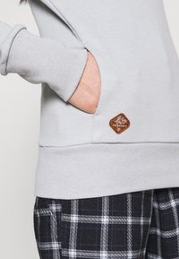 Ljusgrå sweatshirt med ribbade muddar, sidoficka och en logopatch i brunt läder. Tillsammans med mörka rutig byxor med vita linjer.