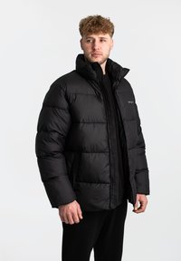 Zwarte pufferjas met een hoge kraag, horizontaal stikselpatroon en ritssluiting. Beschikt over zijzakken en een slank ontwerp.