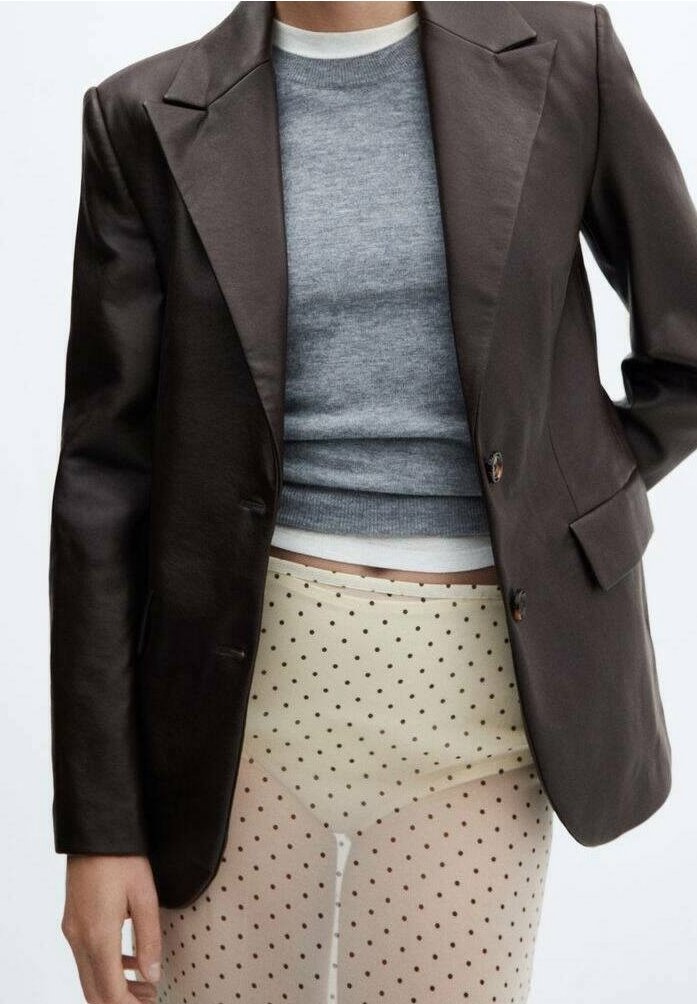 Person, der ein dunkelbraunes Blazer, einen grauen, kurz geschnittenen Strickpullover und durchsichtige beige Strumpfhosen mit schwarzen Punkten trägt.
